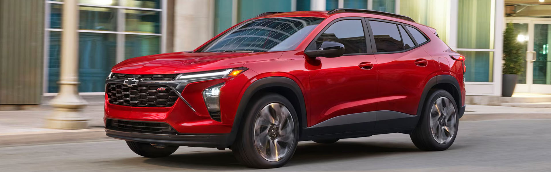 Red 2026 Chevrolet Trax driving through a city street, featuring a bold front grille and motion blur against a modern urban building backdrop.