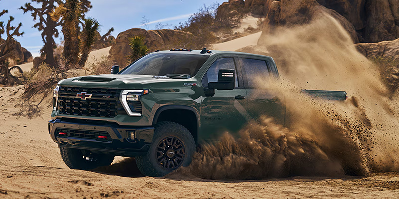 A rugged green pickup truck Silverado-HD powers through sandy desert terrain, kicking up dust. The background features rocky formations and sparse vegetation under a clear sky.