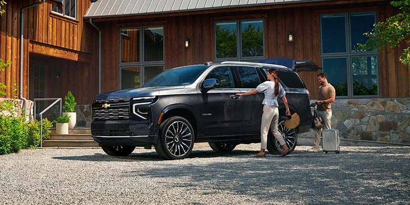 Two people load luggage into the trunk of a black Tahoe parked on a gravel driveway in front of a wood-paneled house, conveying a sense of travel readiness.