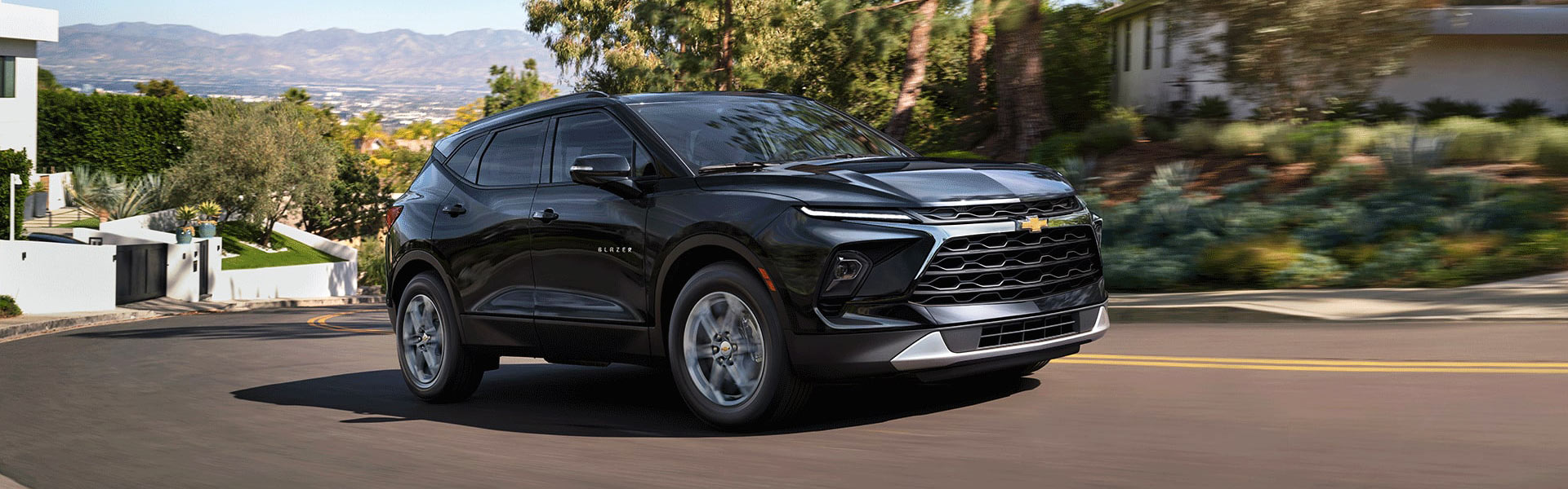 2026 Chevrolet Blazer in black driving on a winding road surrounded by greenery and modern homes.