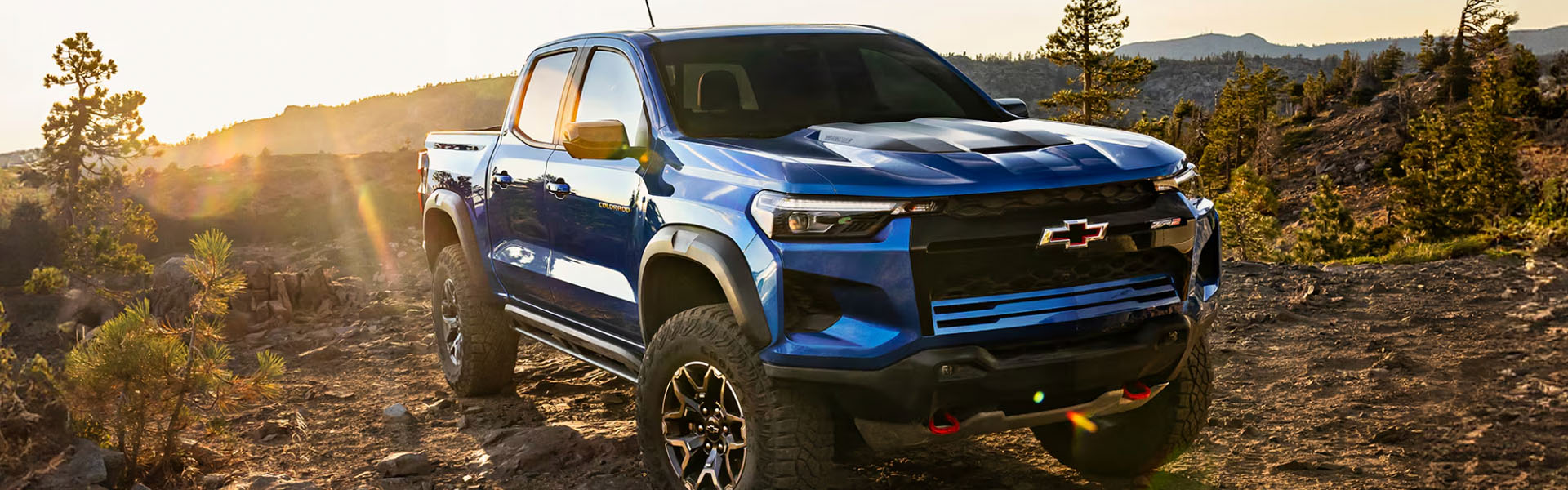 2026 Chevy Colorado ZR2 blue midsize pickup truck tackling rugged off-road terrain at sunset.